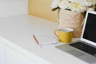 A desk with a laptop, a yellow coffee cup, and a notebook on it. Next to the laptop is a vase filled with white flowers