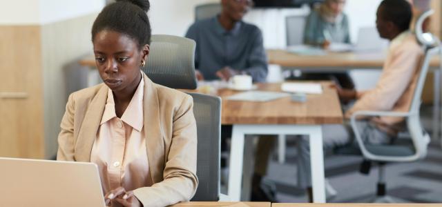 woman works on her laptop in a modern office, with colleagues in the background.